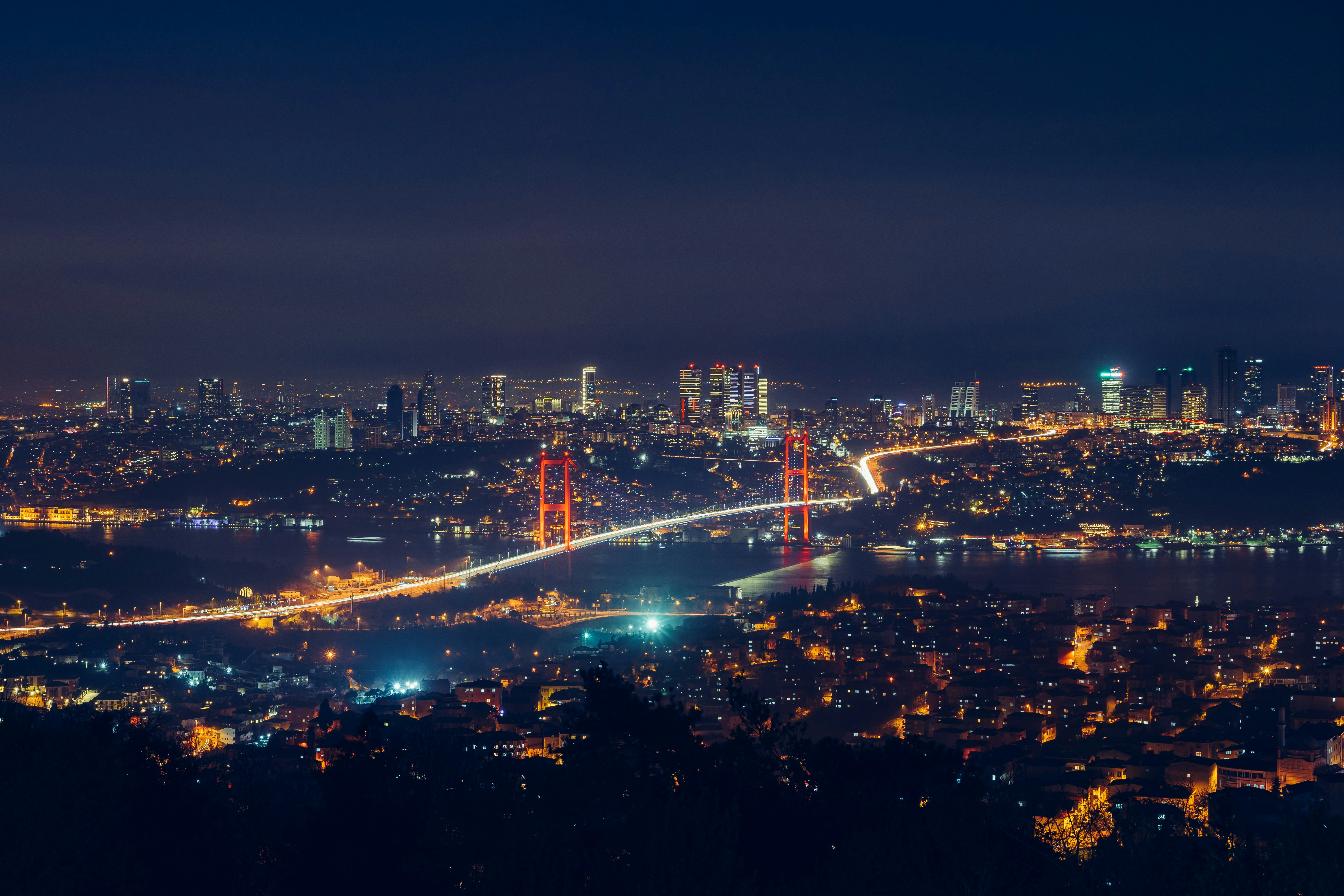 Night view of the Bosphorus in Istanbul, featuring the 15 July Martyrs Bridge illuminated with red lights and the city's vibrant skyline in the background. The bridge shines under the dark night sky, showcasing Istanbul’s lively cityscape.
